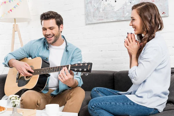 女人坐在沙发上 听男人在家里表演声学吉他 — 图库照片