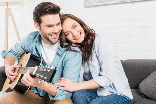 Feliz Casal Sentado Abraçando Sofá Enquanto Homem Tocando Guitarra Acústica — Fotografia de Stock