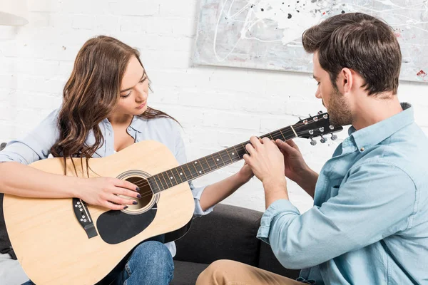 man sitting on couch and teaching woman to play acoustic guitar at home