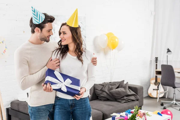 happy couple in party hats looking at each other, embracing and holding birthday present in living room