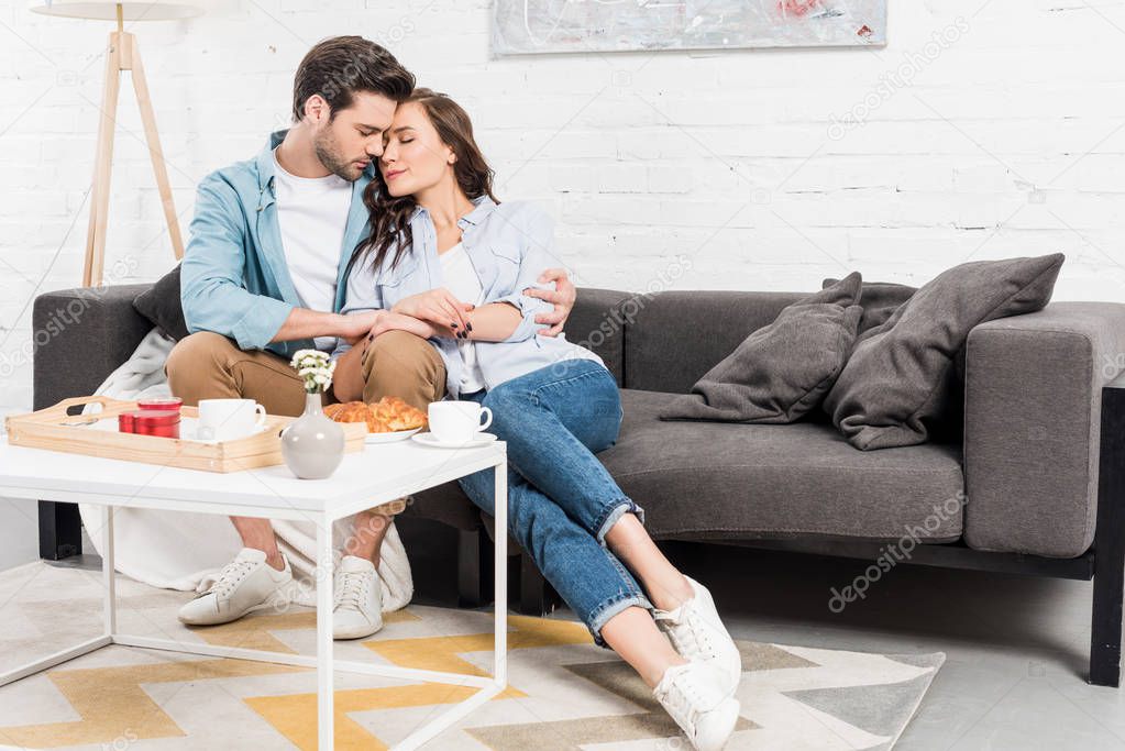 couple sitting on couch and tenderly embracing while having breakfast in living room