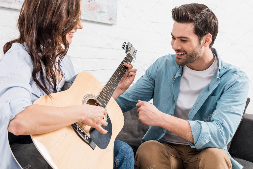 happy couple sitting on couch and playing acoustic guitar at home