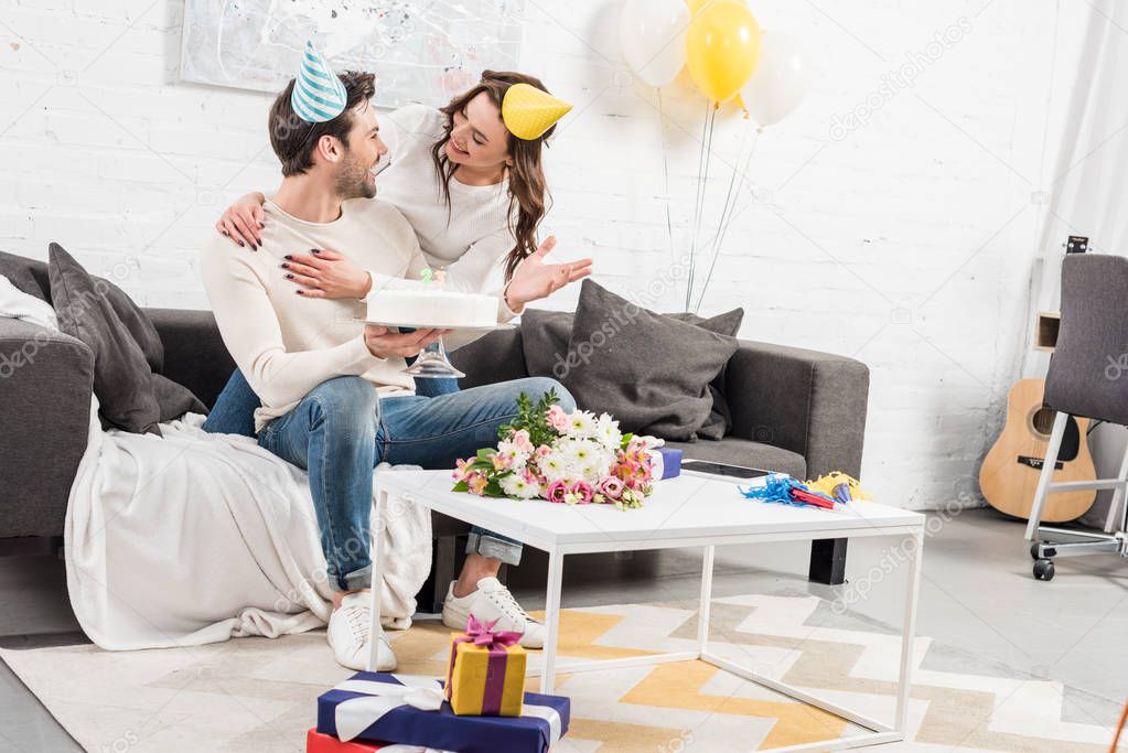 couple in party hats hugging and celebrating birthday with cake in living room