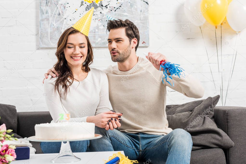 smiling couple in party hats celebrating birthday with cake and party horn at home