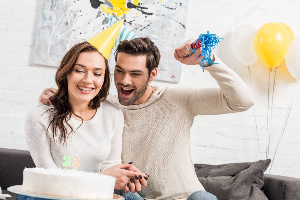 cheerful couple in party hats celebrating birthday with cake and party horn at home