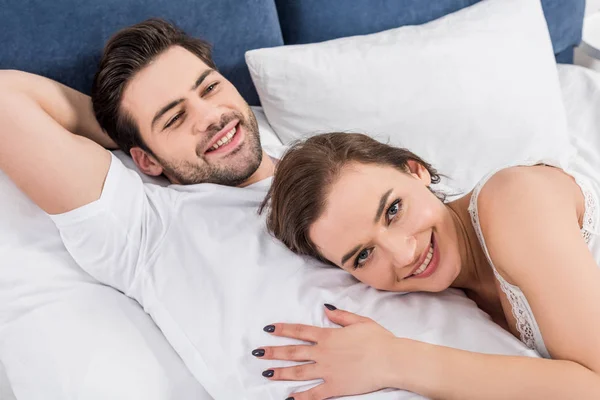 カメラ目線の女性ながら自宅のベッドで寄り添う笑顔のカップル — ストック写真