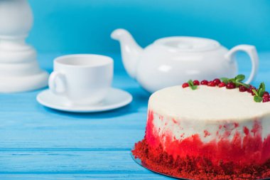 cake decorated with red currants and mint leaves between white cup, tea pot and stand isolated on blue clipart