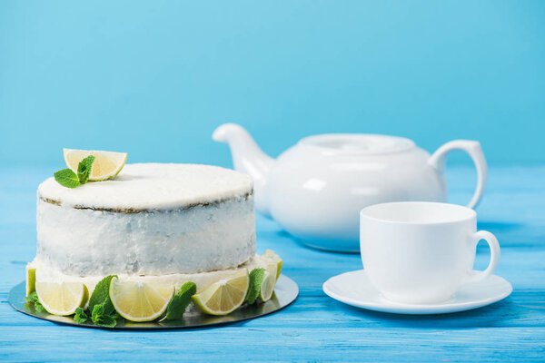 cake decorated with slices of lime near white cup and tea pot isolated on blue