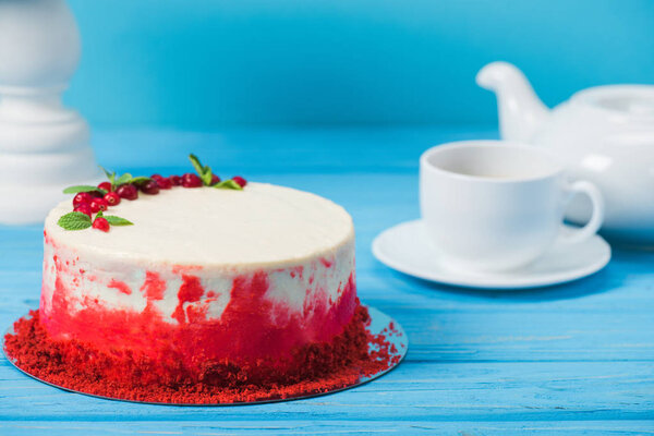 cake decorated with red currants and mint leaves between white cup, tea pot and stand isolated on blue