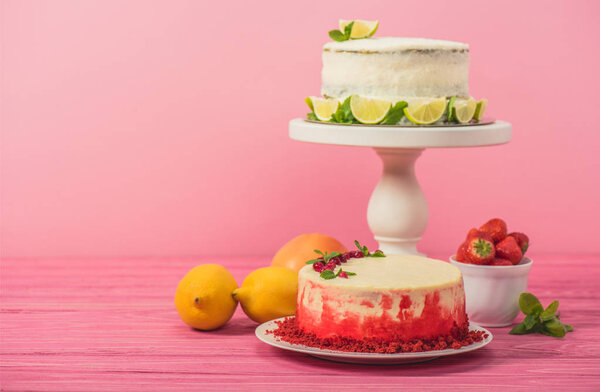 cake decorated with currants and mint leaves near fruits and white cake with lemon slices on pink wooden surface isolated on pink