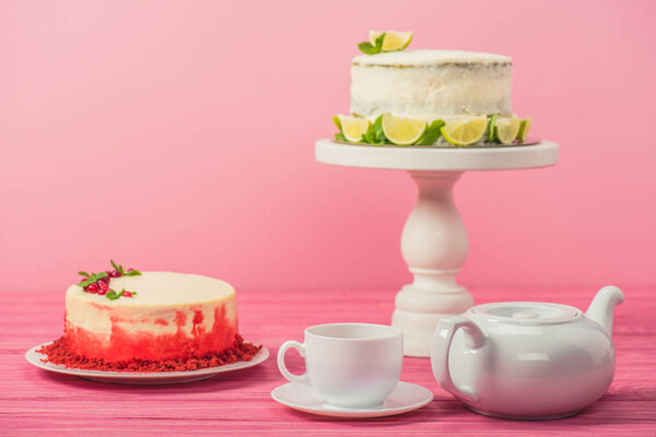cakes decorated with currants, mint leaves and lime slices near cup and tea pot isolated on pink