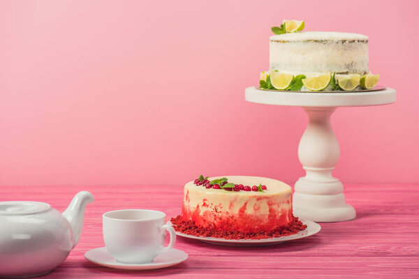 cakes decorated with currants, mint leaves and lime slices near cup and tea pot on wooden surface isolated on pink