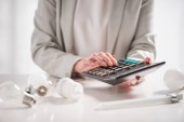 cropped view of woman using calculator near lamps on white background, energy efficiency concept