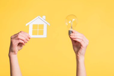 cropped view of female hands holding paper house and led lamp in hands isolated on yellow, energy efficiency at home concept clipart