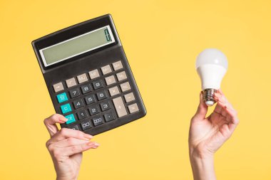 cropped view of woman holding fluorescent lamp and calculator in hands isolated on yellow, energy efficiency concept  clipart