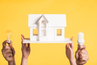 cropped view of woman holding carton house model near man with led and fluorescent lamps isolated on yellow, energy efficiency at home concept