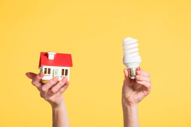 cropped view of woman holding fluorescent lamp and house model in hands isolated on yellow, energy efficiency at home concept clipart