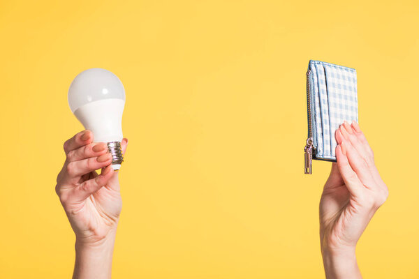 cropped view of woman holding fluorescent lamp and wallet in hands isolated on yellow, energy efficiency concept