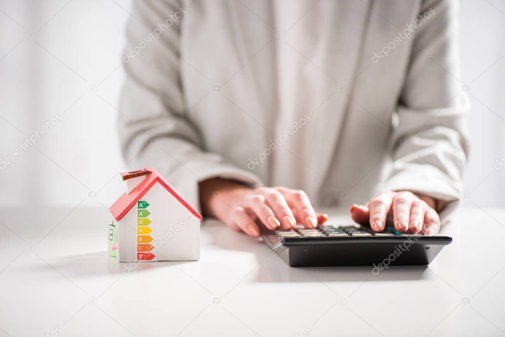 cropped view of woman using calculator near carton house on white background, energy efficiency at home concept