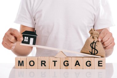 partial view of man holding moneybag and paper house on scales on cubes with mortgage lettering isolated on white clipart
