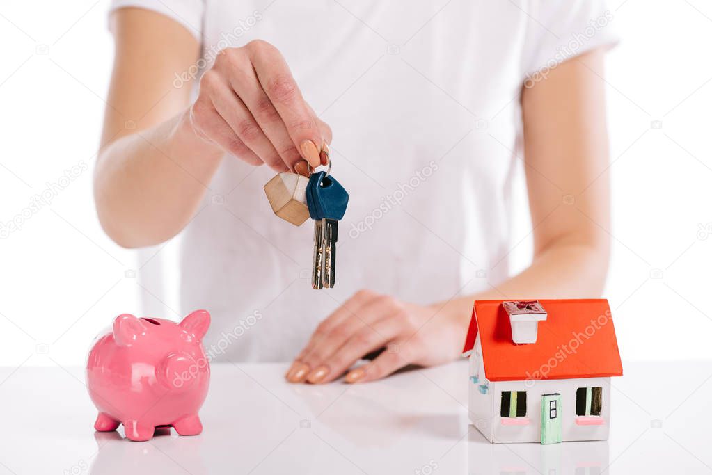 partial view of woman holding keys near house model and piggy bank isolated on white, mortgage concept