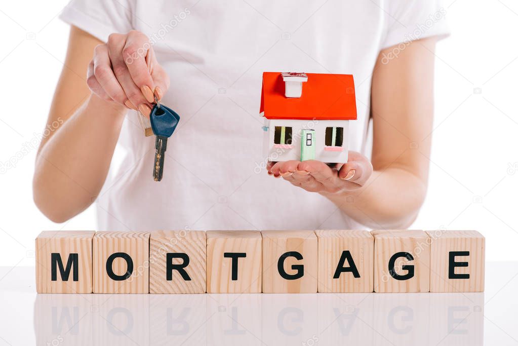 partial view of woman holding house model and keys near wooden cubes with mortgage lettering isolated on white