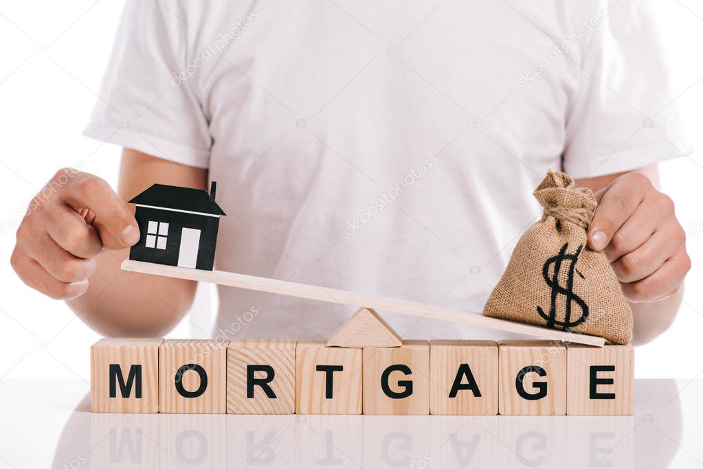 partial view of man holding moneybag and paper house on scales on cubes with mortgage lettering isolated on white