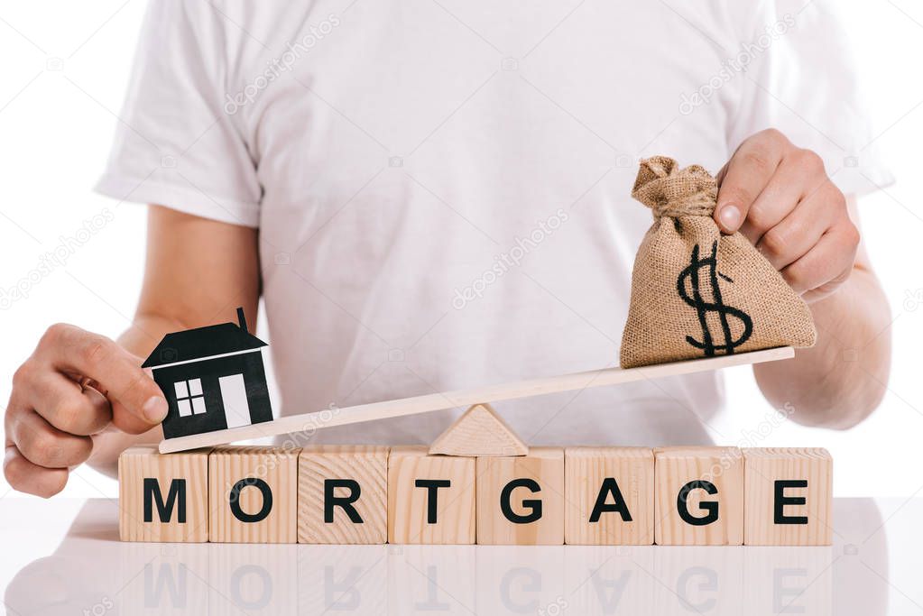 cropped view of man holding moneybag and paper house on scales on cubes with mortgage lettering isolated on white