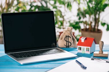 selective focus of pen, clipboard, laptop with blank screen near stamp, house model and moneybag with dollar sign on wooden desk, mortgage concept clipart