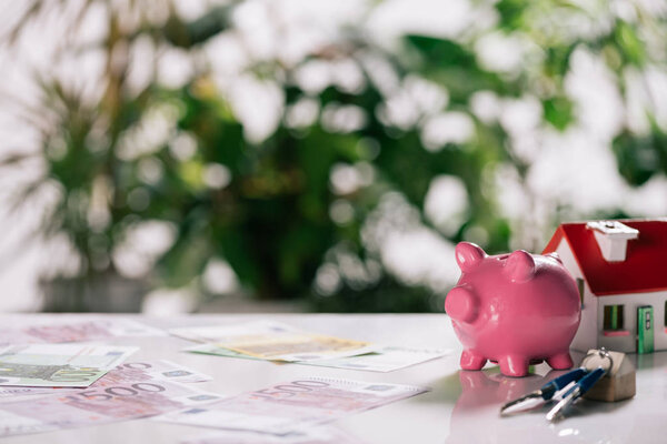 selective focus of euro banknotes, keys, piggy bank and house model on white desk, mortgage concept