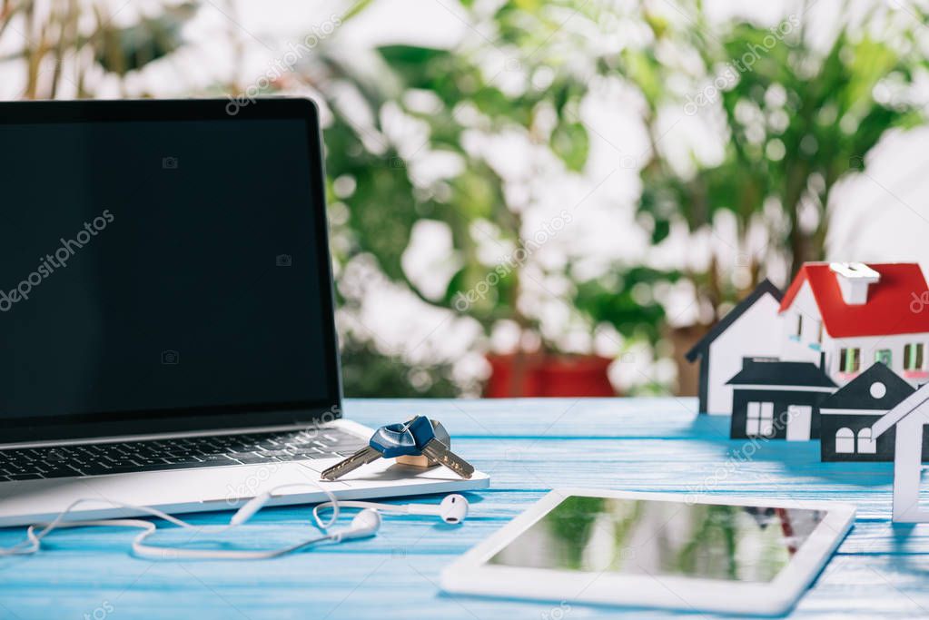 selective focus of digital tablet, laptop with blank screen and keys near headphones and house model on wooden desk, mortgage concept