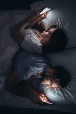 young couple lying back to back in bed and using smartphones at night clipart
