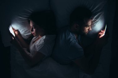couple lying back to back in bed and using smartphones at night clipart