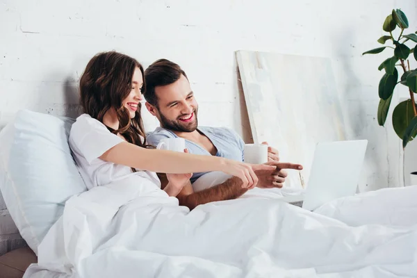 成人夫妇放松 躺在柔软的白色床上用品和手指指向笔记本电脑 — 图库照片