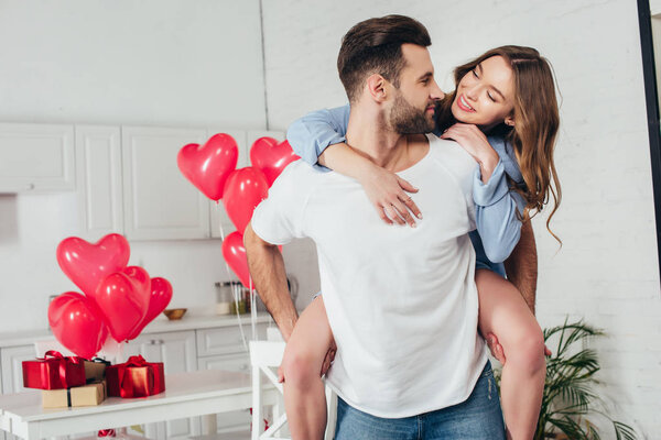 young man giving piggyback ride to smiling girlfriend at home with st valentine day decoration 