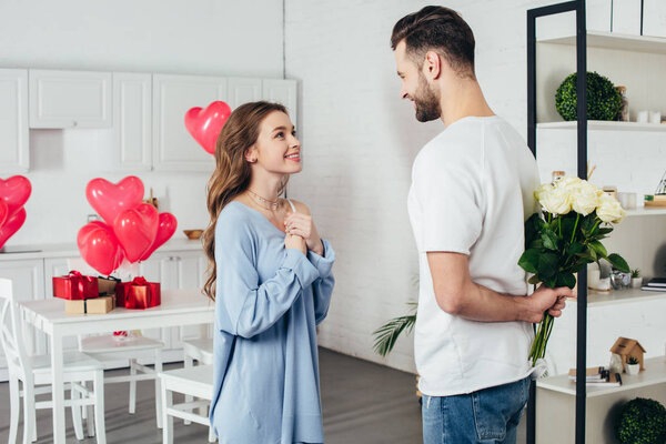 young smiling girl in joyful expectation of st valentines day gift while smiling boyfriend holding roses bouquet behind back 