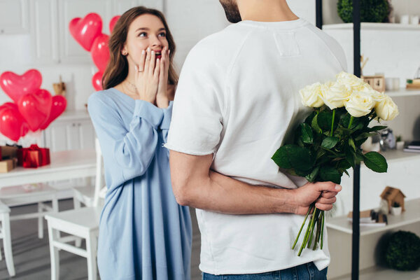 partial view of a young man holding bouquet of flowers behind back while smiling girlfriend waiting for surprise