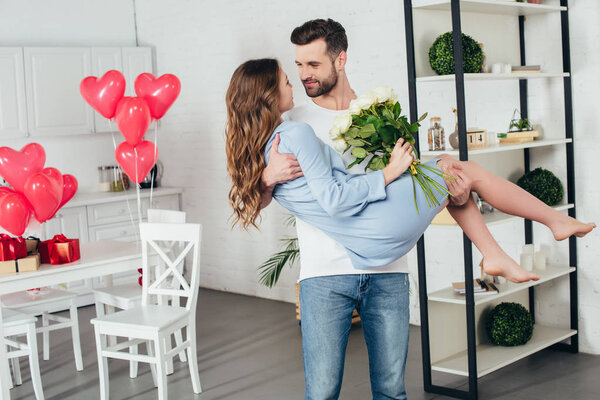 young man holding smiling girlfriend with roses bouquet in arms at st valentine day