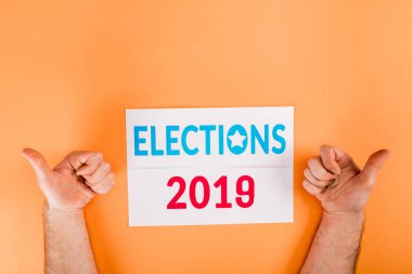cropped view of man showing thumbs up near card with 'elections 2019' lettering isolated on orange clipart