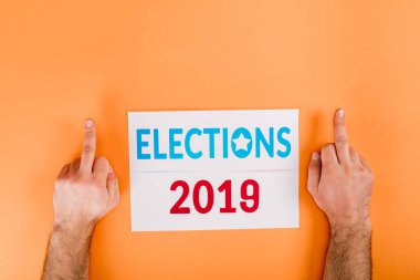 cropped view of man showing middle fingers near card with 'elections 2019' lettering isolated on orange