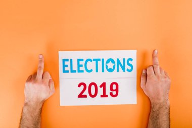 cropped view of man showing middle fingers near card with 'elections 2019' lettering isolated on orange clipart