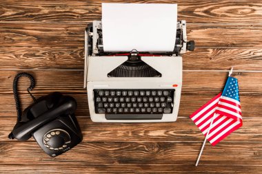 top view of typewriter, rotary dial telephone and american flag on wooden table