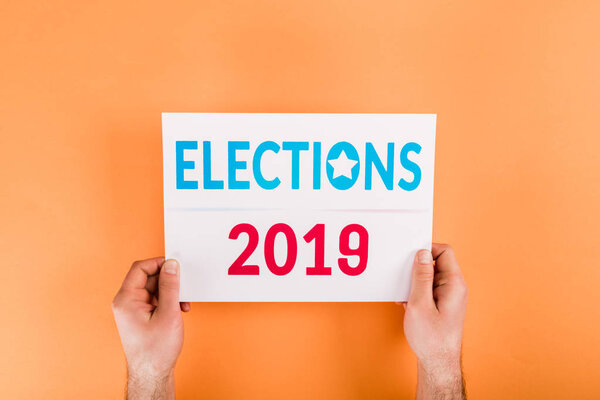 cropped view of man holding card with 'elections 2019' lettering isolated on orange