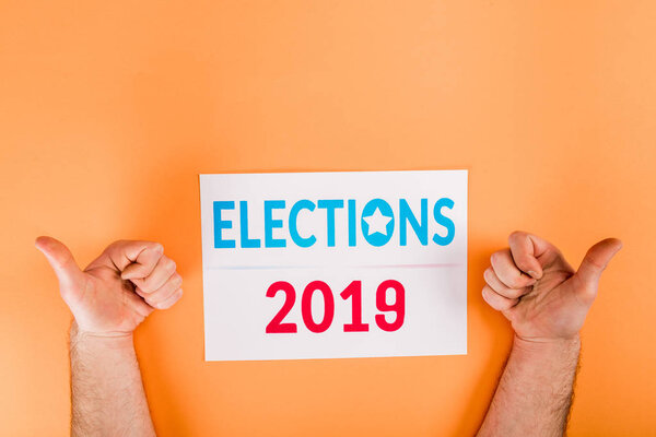 cropped view of man showing thumbs up near card with 'elections 2019' lettering isolated on orange