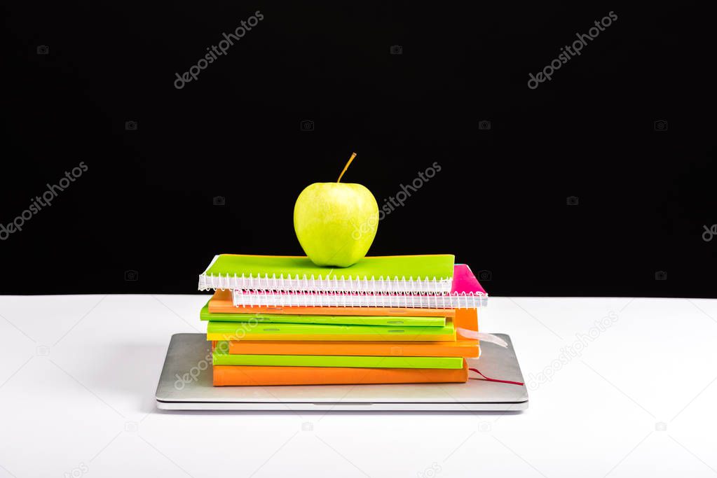 colorful notebooks, apple and laptop on desk isolated on black