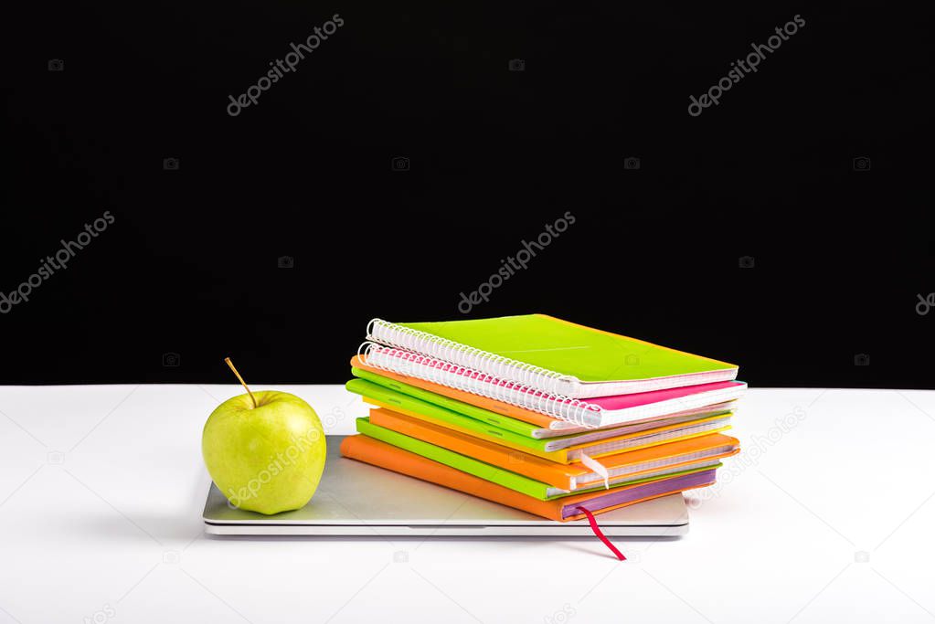 colorful notebooks, apple and laptop on desk isolated on black with copy space