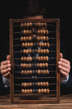 businessman holding abacus and looking at camera while sitting at wooden table clipart
