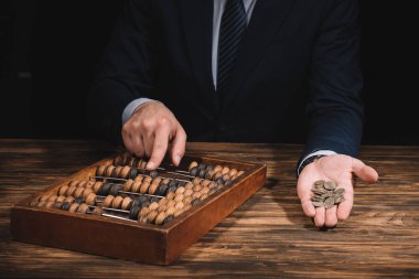 cropped shot of businessman holding coins and using abacus at wooden table clipart