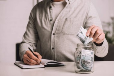 cropped shot of man writing in notebook and putting russian rubles banknotes in glass jar clipart