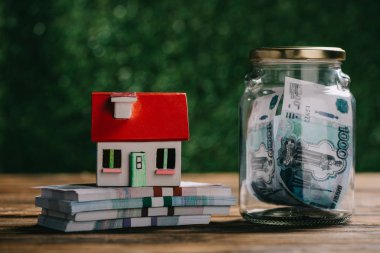 close-up view of glass jar with money, house model and rubles banknotes on wooden table clipart